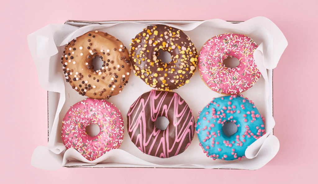 Sechs bunte Donuts mit Glasur und Streuseln in einer weißen Box auf rosafarbenem Hintergrund
